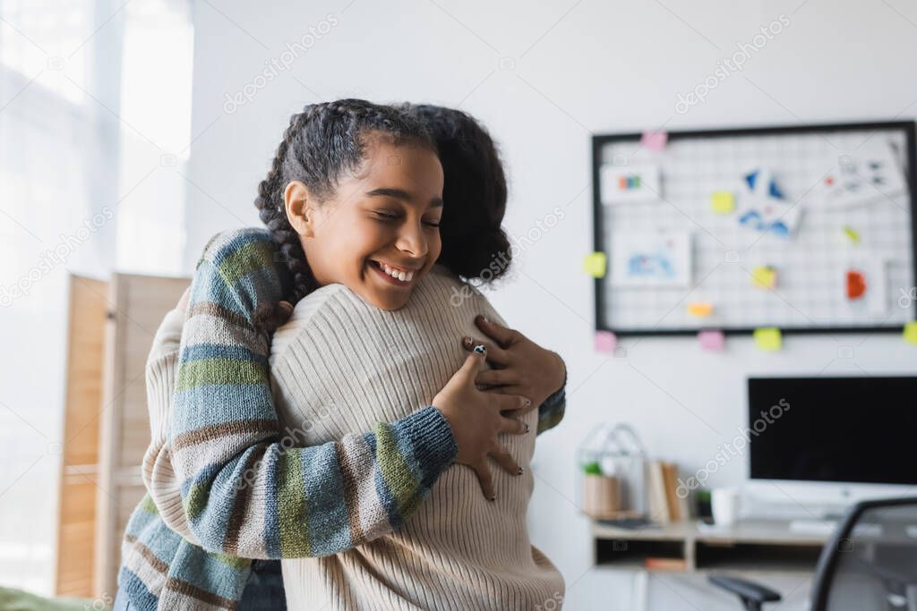 smiling african american teenage girl with closed eyes hugging mom at home