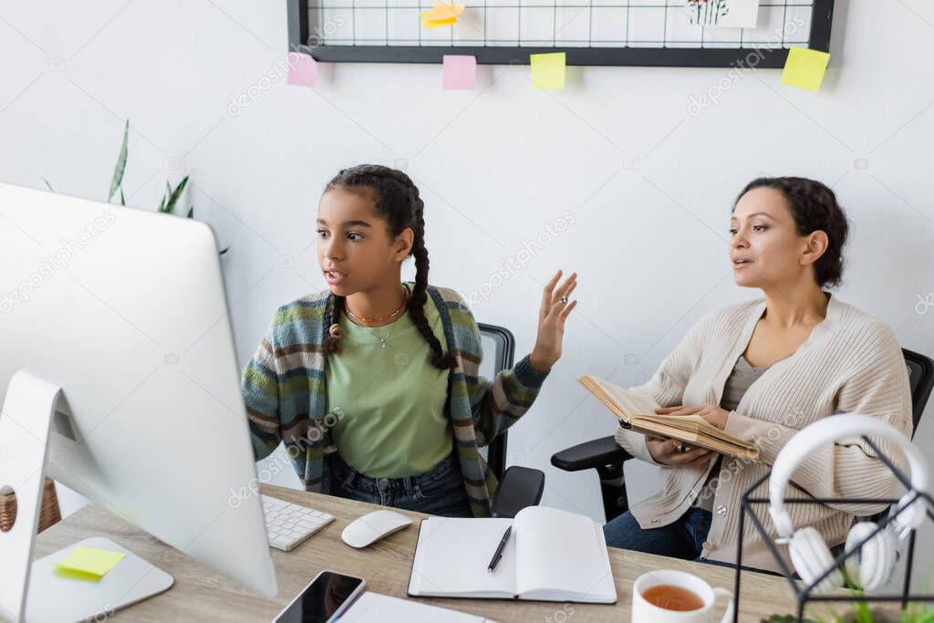 african american girl gesturing while looking at computer monitor near mother with book