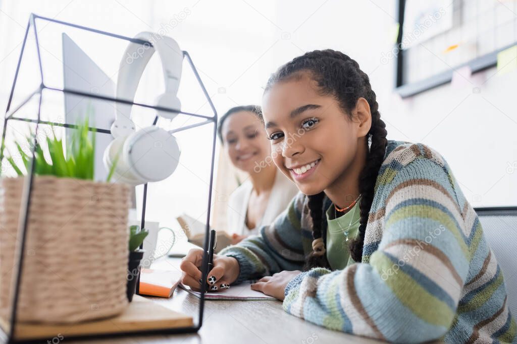 happy african american teen girl looking at camera writing in notebook near blurred mom with book