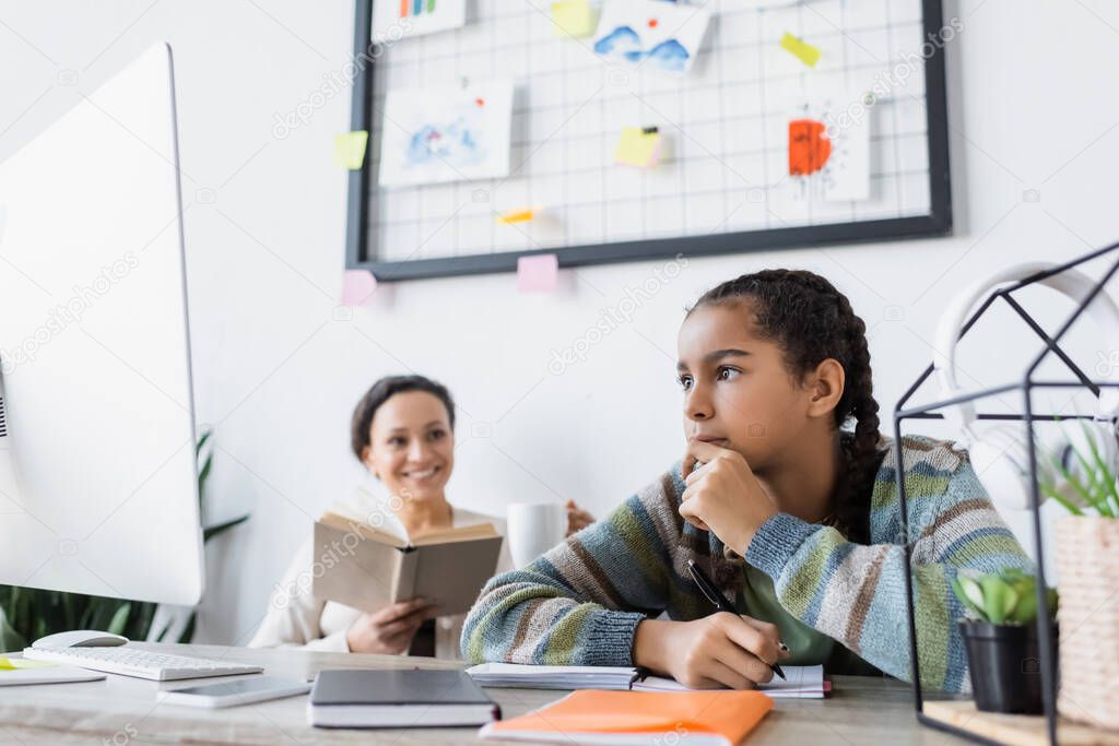 thoughtful african american girl doing homework near laptop and mom with book smiling on blurred background