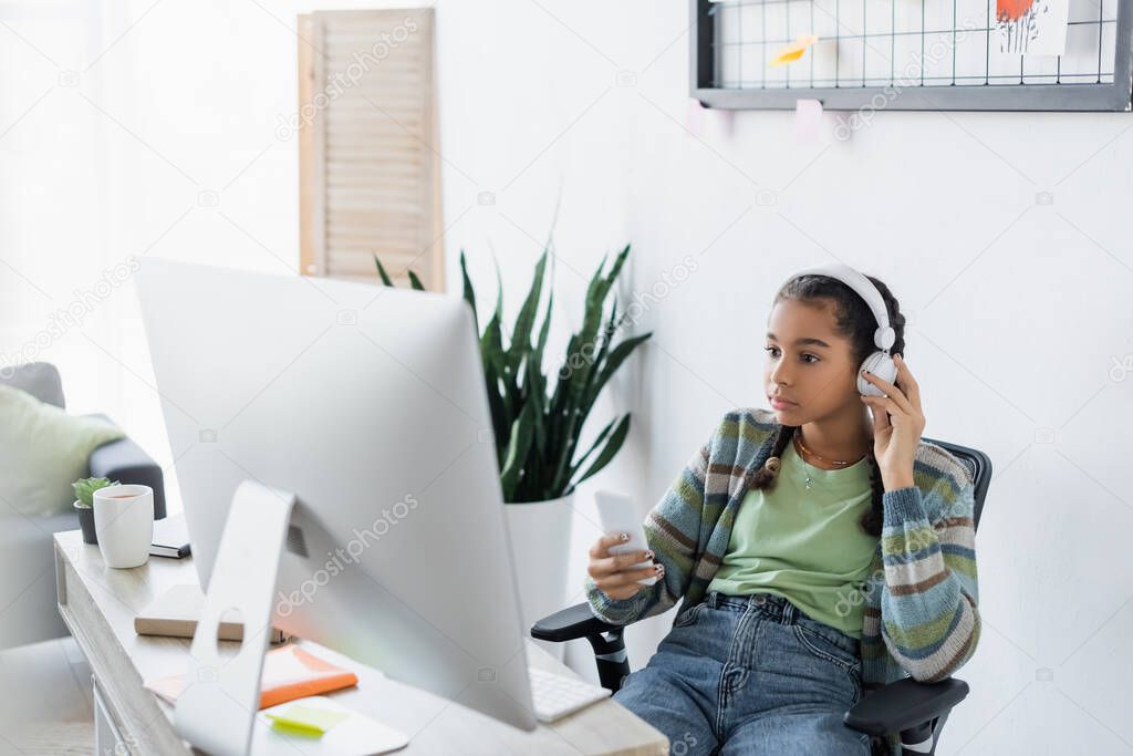 african american teen girl holding cellphone while sitting near computer monitor