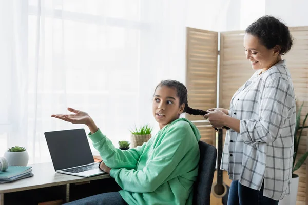 african american girl pointing at laptop with blank screen while mother braiding her hair