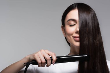 pleased brunette woman using hair straightener isolated on grey clipart