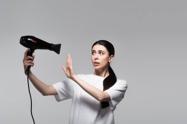 careful woman in white t-shirt holding hair dryer isolated on grey clipart