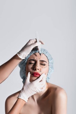 Plastik eldivenli estetik cerrah cerrahi şapkalı hastanın yanında neşter tutuyor ve yüzünde gri çizgiler var.