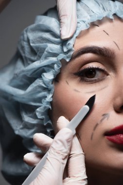 close up of plastic surgeon in latex gloves holding scalpel near woman with marked lines on face isolated on dark grey clipart