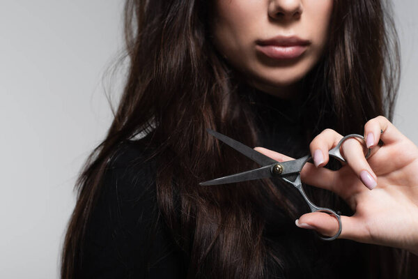 cropped view of young woman in black turtleneck holding scissors near long hair isolated on grey