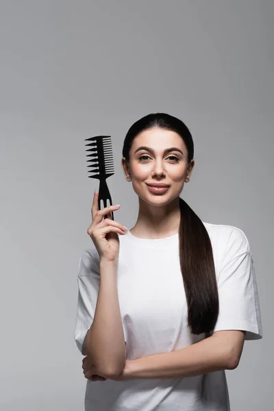 happy woman in white t-shirt holding comb near straight hair isolated on grey