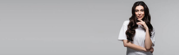 young cheerful woman with long wavy hair looking at camera isolated on grey, banner