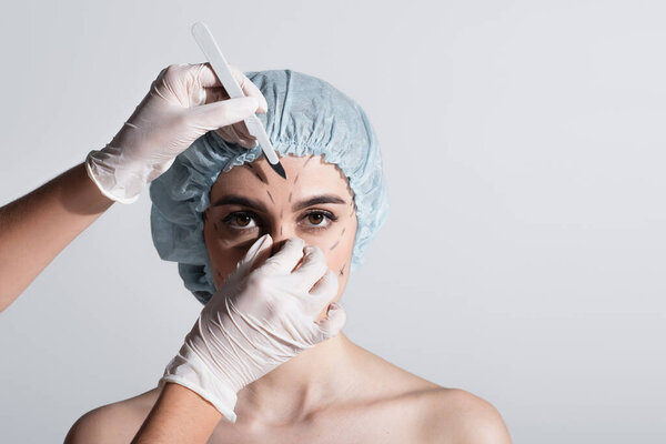 plastic surgeon in latex gloves holding scalpel near woman in surgical cap with marked lines on face isolated on grey