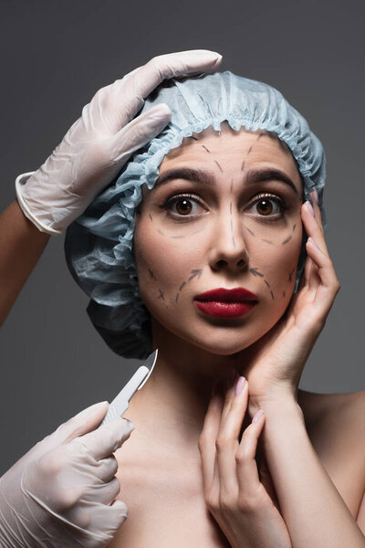 plastic surgeon in latex gloves holding scalpel near scared woman with marked lines on face isolated on dark grey