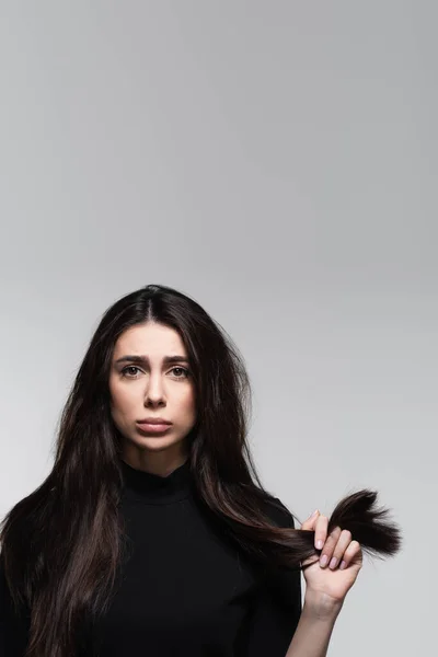 sad young woman in black turtleneck holding damaged hair isolated on grey