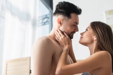 happy young lovers looking at each other while standing face to face in bedroom