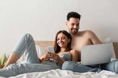 pleased couple smiling while using gadgets in bedroom at home