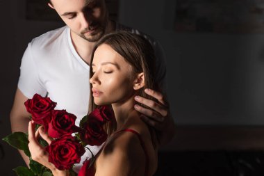 sensual woman with closed eyes holding red roses near man in white t-shirt in dark bedroom