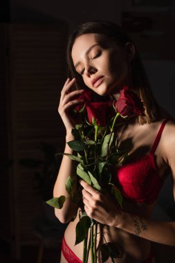 passionate woman in red underwear standing with closed eyes and roses on dark background