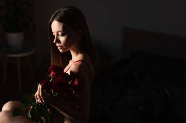 seductive woman sitting in dark bedroom with bouquet of red roses