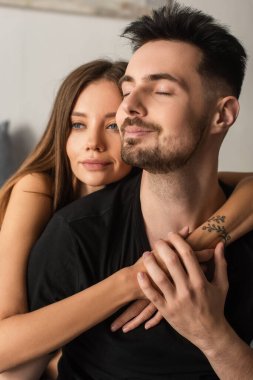 happy man with closed eyes touching hands of tender woman hugging him in bedroom