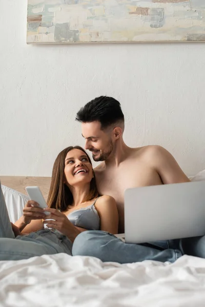 Happy Young Couple Looking Each Other While Using Gadgets Bed — 스톡 사진