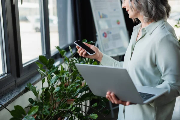 オフィスの植物の近くに空白の画面とラップトップを持つスマートフォンを保持している実業家の作物ビュー — ストック写真