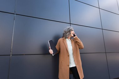 Şehir caddesinde cep telefonuyla konuşurken elinde kağıt klasör tutan palto ve gözlüklü pozitif iş kadını. 