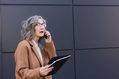 Kentsel caddede akıllı telefondan konuşurken elinde kağıt klasör olan gri saçlı bir yönetici. 