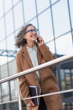 Palto giymiş neşeli bir iş kadını elinde belgelerle dışarıda parmaklıkların yanında cep telefonuyla konuşuyor. 
