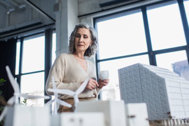 Elinde kahve fincanı olan gri saçlı iş kadını ofisteki binaların modellerine bakıyor. 