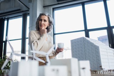 Pensive businesswoman holding cup near blurred models of buildings in office  clipart