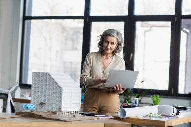 Ofisteki bina ve planların yanında dizüstü bilgisayar kullanan orta yaşlı bir iş kadını. 