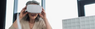 Grey haired businesswoman using virtual reality headset near blurred model of building in office, banner  clipart