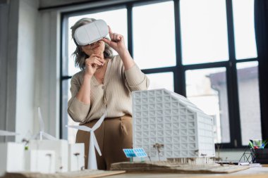 VR kulaklıklı gri saçlı iş kadını ofisteki binaların modellerinin yanında duruyor. 