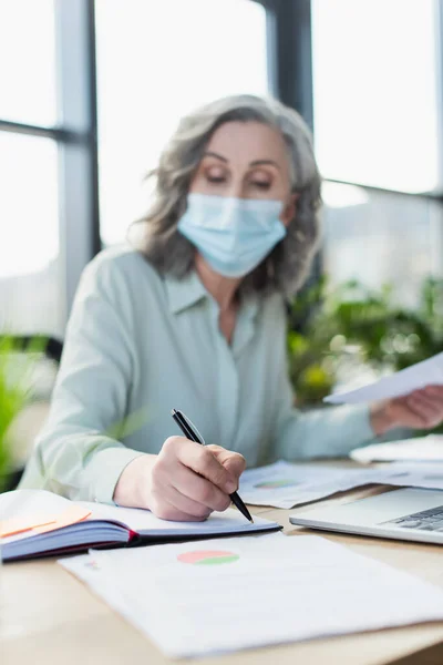 Verschwommene Geschäftsfrau Medizinischer Maske Schreibt Auf Notizbuch Neben Laptop Büro — Stockfoto