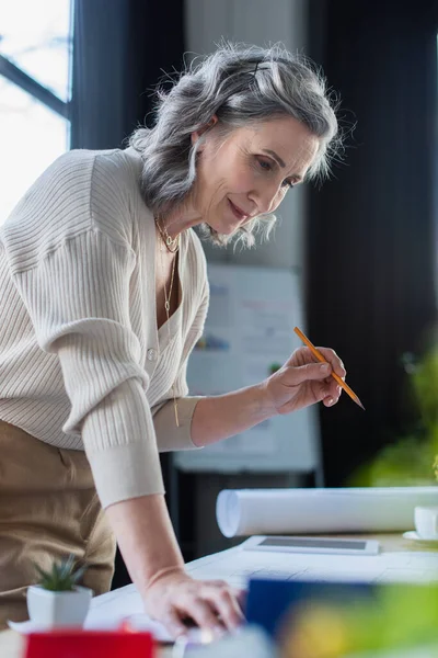 Mature businesswoman holding pencil near blueprint and digital tablet in office