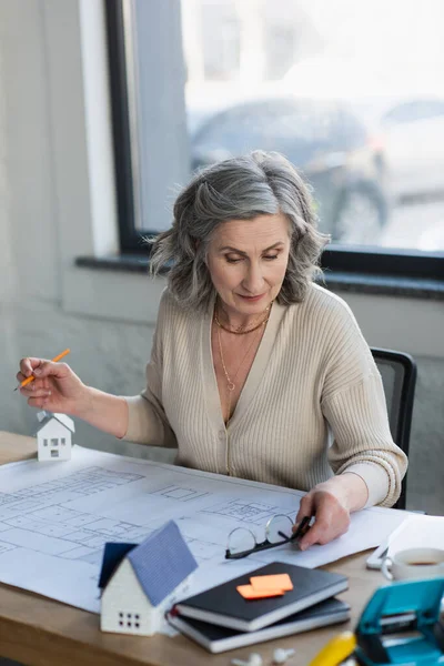 Businesswoman taking eyeglasses near blueprint and models of houses in office