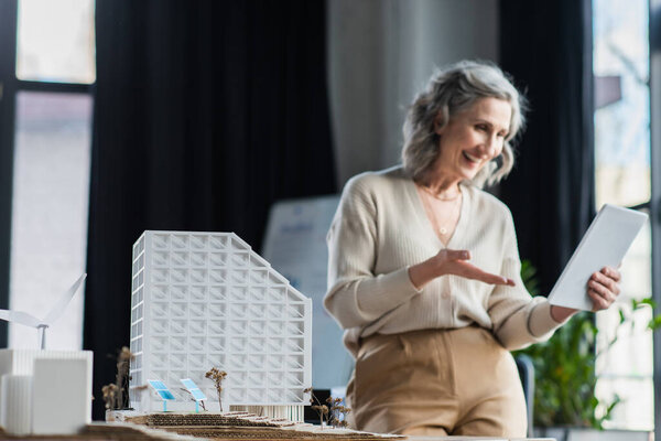 Models of buildings near blurred businesswoman having video call on digital tablet in office 