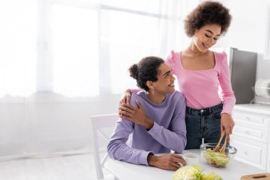 Afrikalı Amerikalı kadın erkek arkadaşına sarılıyor ve mutfakta salata pişiriyor. 