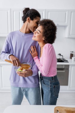 Smiling african american man holding bowl of salad and whispering to girlfriend in kitchen  clipart