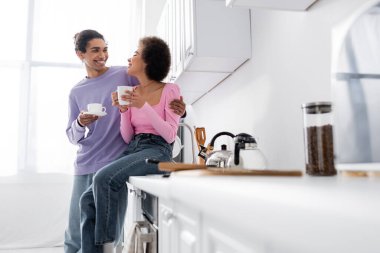 Genç Afro-Amerikalı bir adam mutfakta elinde fincanla kız arkadaşına sarılıyor. 