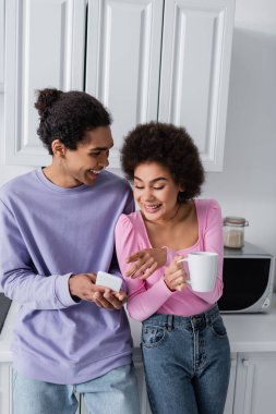 Evdeki erkek arkadaşının yanındaki akıllı telefonu işaret eden fincanlı Afrikalı Amerikalı kadın. 