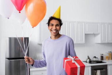 Evde kameraya bakarken elinde hediye ve balon tutan mutlu Afro-Amerikan adam. 