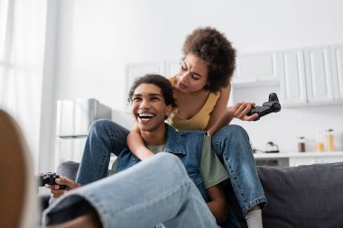 KYIV, UKRAINE - 9 Kasım 2021: hoşnutsuz Afro-Amerikan kadın kanepede neşeli erkek arkadaşının yanında joystick tutuyor 