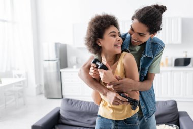 KYIV, UKRAINE - 9 Kasım 2021: gülümseyen Afro-Amerikan erkek evinde kız arkadaşını joystick ile kucaklıyor 