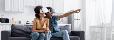 Heyecanlı Afro-Amerikan adam, VR kulaklıklı, parmaklarıyla evde gülümseyen kız arkadaşını işaret ediyor. 