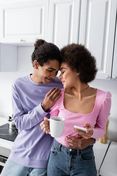 Young African American Man Hugging Girlfriend Cup Cellphone Kitchen — Stock Photo, Image