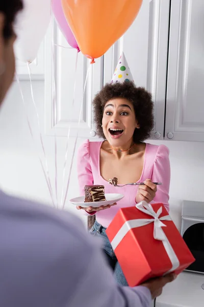 Aufgeregte Afroamerikanerin Hält Kuchen Neben Verschwommenem Freund Mit Geschenk Und — Stockfoto