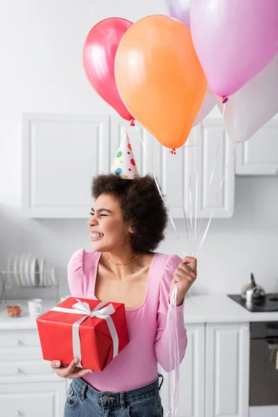 幸せなアフリカ系アメリカ人女性でパーティーキャップ保持ギフトと風船でキッチンに自宅 — ストック写真