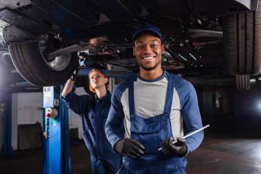Smiling african american mechanic with digital tablet looking at camera near colleague and car in garage  clipart