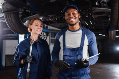 Positive multiethnic mechanics holding wrench and digital tablet near car in garage  clipart