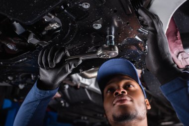 Low angle view of african american mechanic in cap using wrench while working in car service  clipart